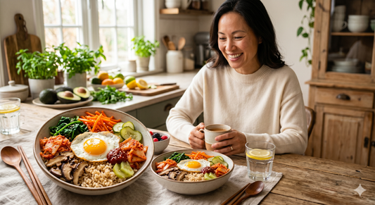 Frau sitzt lächelnd vor zwei Bibimbap-Bowls mit buntem Gemüse, Ei und Kimchi an einem Holztisch